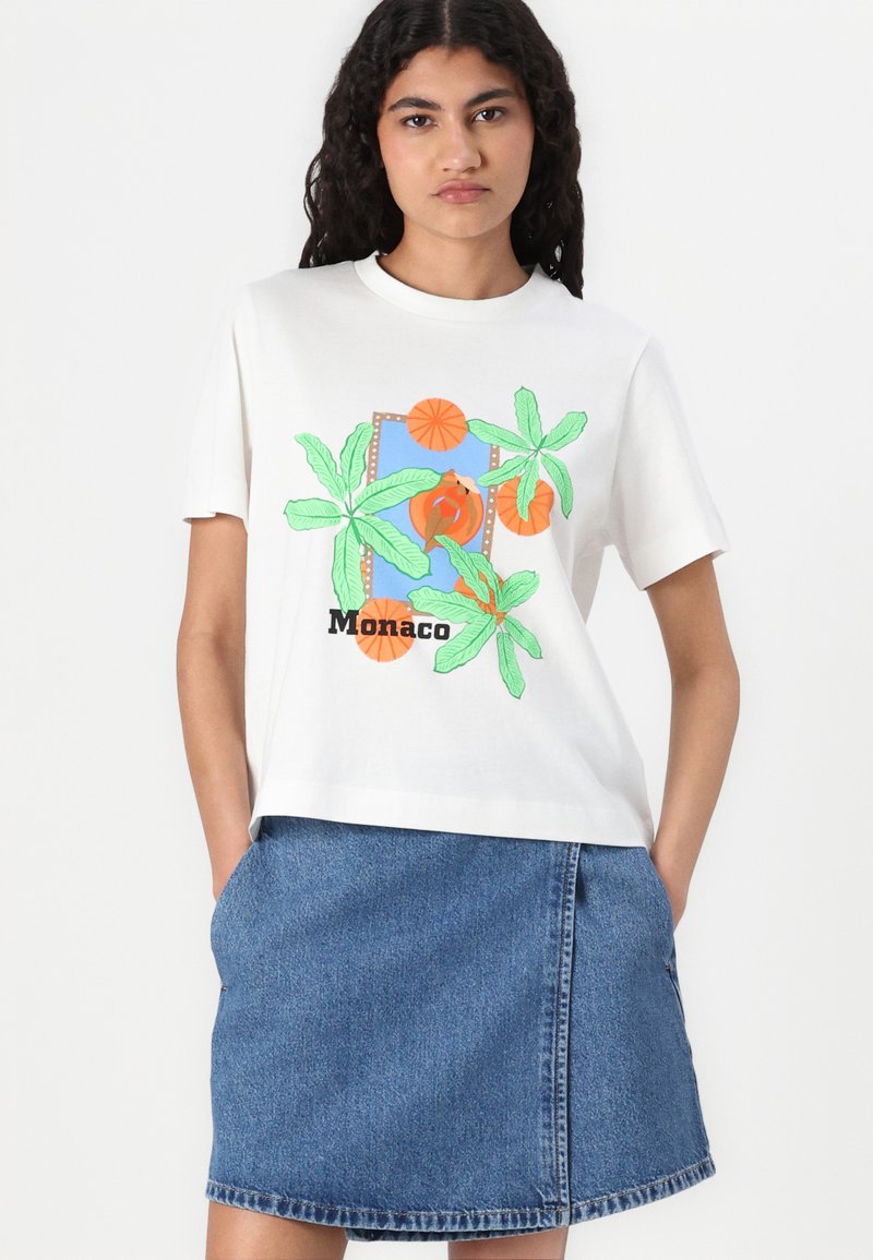 T-shirt blanc avec un imprimé graphique coloré de palmier, portant le texte "Monaco" ; associé à une jupe en denim. Manches courtes, coupe décontractée.