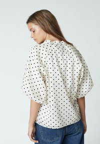Frau trägt eine weiße Bluse mit schwarzen Punkten und Puffärmeln, von hinten gezeigt, kombiniert mit blauen Jeans.