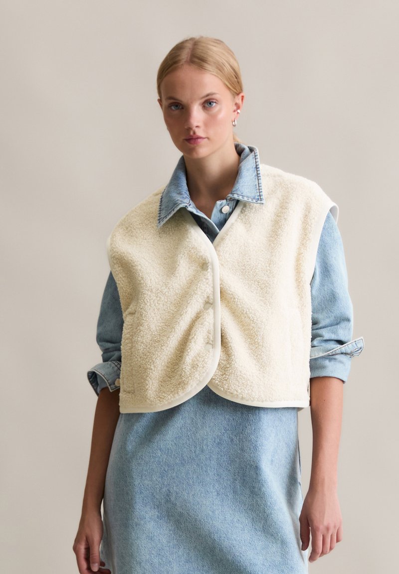 Crème-kleurige, gestructureerde fleece vest met een ronde vorm, knopen aan de voorkant en een spijkerblouse eronder. Draagt over een blauwe spijkerjurk.
