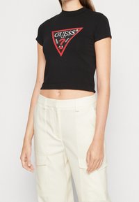 Schwarzes, cropped T-Shirt mit taillierter Passform, das ein rotes und weißes dreieckiges Logo auf der Vorderseite zeigt. Glattes Baumwollmaterial mit hohem Kragen.