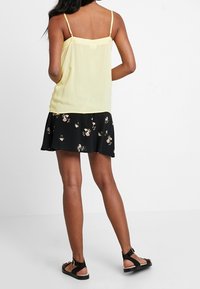 Žena stojící zády k fotoaparátu, oblečená v žlutém tílku bez rukávů, černé květinové sukni a černých sandálech na bílém pozadí.