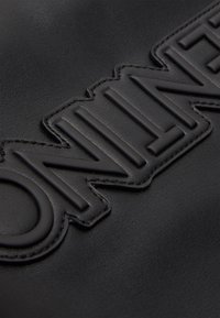 Musta nahkalaukku, jossa on kohotettu kirjonta, sileä pinta ja erottuva ommel. Läheinen lähikuva materiaalista ja logon yksityiskohdista.