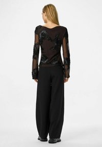 Must pikkade varrukatega top läbipaistvast kangast, millel on keerukas must lilleline tikand. Sobitatud mustade laiade jalgadega pükste ja tossudega.