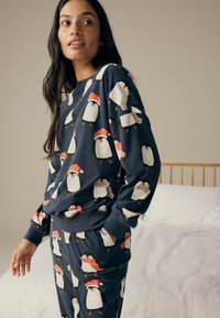Marineblaues Schlafanzugset mit Pinguinmustern, die Weihnachtsmützen tragen. Weiches Material mit gerippten Bündchen und Taschen an der Hose, lässiger Schnitt.