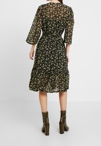 Robe noire transparente à fleurs jaunes, manches trois-quarts, taille cintrée, ourlet volanté. Associée à des bottines brunes à motifs.