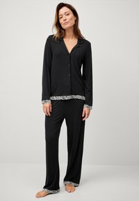 Sæt bestående af sorte pyjamas, der har en knappet skjorte med krave og lange ærmer, som er kombineret med løstsiddende bukser, begge prydet med pyntelace.