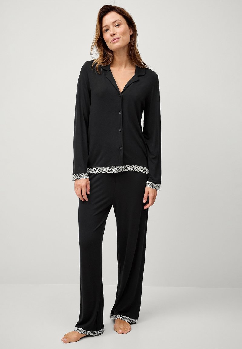 Sæt bestående af sorte pyjamas, der har en knappet skjorte med krave og lange ærmer, som er kombineret med løstsiddende bukser, begge prydet med pyntelace.