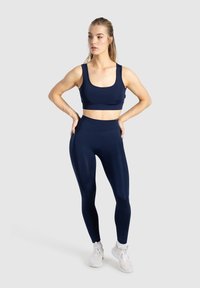 Námornická sportovní podprsenka a legíny s vysokým pasem vyrobené z hladké, elastické tkaniny. Podprsenka má široké ramínka. Modelka stojí v uvolněné póze.