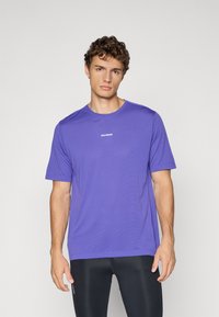 Ung mand med krøllet hår iført en lilla Salomon T-shirt og sorte træningsleggings, der står foran en ensfarvet baggrund.