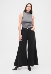 Top cinzento sem mangas com gola alta combinado com calças pretas de perna larga e cintura alta. As calças têm uma textura lisa e bolsos laterais. Botas pretas de salto.