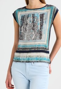 Printed lightweight short-sleeve top featuring a square design with blue, black, and white floral patterns and navy trim, paired with light jeans.