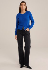 Zils adītais džemperis ar apaļu kakla izgriezumu un pogu aizdari, apvienots ar melniem platām kājām džinsiem un melnām smailām kurpēm. Gluda tekstūra.