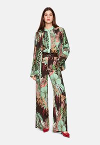 Helle Weitbein-Hosen und passendes Hemd aus einem dunkelbraunen Stoff mit lebhaften floralen Mustern in Grün, Rot und Pfirsich. Inklusive einem braunen Gürtel.