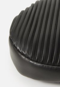 Cuscino in pelle nera con texture a righe verticali. Forma arrotondata, superficie liscia e perimetro leggermente rialzato.