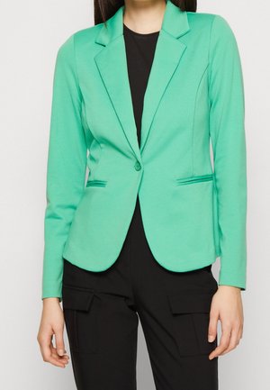 Femme portant un blazer vert menthe cintré sur un top noir et un pantalon cargo noir, debout devant un fond clair uni.