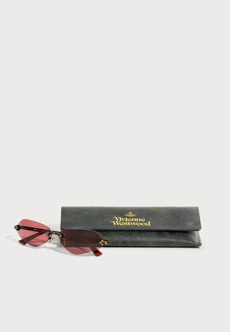 Lunettes de soleil sans monture avec verres roses et accents dorés placées à côté d'un étui noir Vivienne Westwood avec inscriptions dorées.