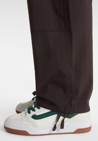 White sneakers with green accents, featuring a textured leather upper and brown rubber sole, paired with dark brown textured pants.