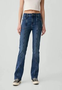 Ženska, oblečena v temno modre naravne jeans hlače s prednjimi šivi, v kombinaciji s črno-belimi športnimi copati in belim skrajšanim zgornjim delom.