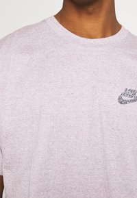 Světle růžové tričko s krátkým rukávem vyrobené z měkké látky, které má texturované, vícolorované logo Nike na levé straně hrudi.