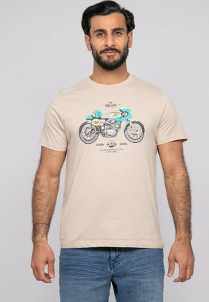 Homme portant un t-shirt beige avec un graphisme vintage de moto turquoise et blanc, et un jean bleu, sur un fond uni.