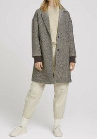 Cappotto grigio a fantasia pied de poule con colletto a revers, polsini viola e vestibilità comoda, abbinato a pantaloni beige e scarpe chiare.