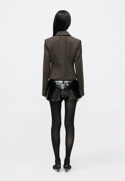 Testre szabott barna csíkos blézer, mely megfelelően illeszkedik, fényes fekete vinil sorttal és fekete áttetsző harisnyával párosítva, hátulról nézve.