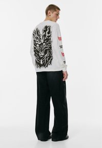 Junger Mann in einem weißen Langarmshirt mit großem schwarzen Monstergesicht-Print auf dem Rücken, roten und schwarzen Schriftzügen auf den Ärmeln und einer schwarzen weiten Hose.