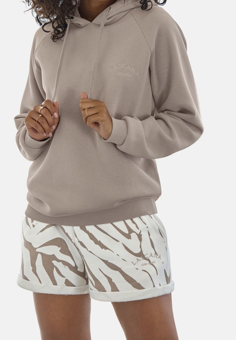 Femme portant un sweat à capuche beige et un short blanc avec des rayures zébrées marron, tous deux avec le logo "Lascana", les mains tenant les cordons du sweat à capuche.