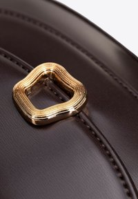 Gros plan sur un sac en cuir marron présentant une surface texturée et une boucle rectangulaire dorée ondulée avec de fines lignes et des détails de couture.