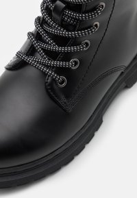 Black leather ankle boot with round toe, textured surface, and contrasting white and black laces. Features metallic eyelets and rubber sole.