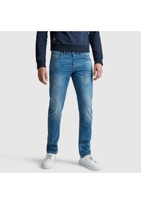 Jeans de mezclilla azul con un corte ajustado, con áreas desgastadas, cinco bolsillos y una clásica tapeta de botones. Combinados con zapatillas blancas.