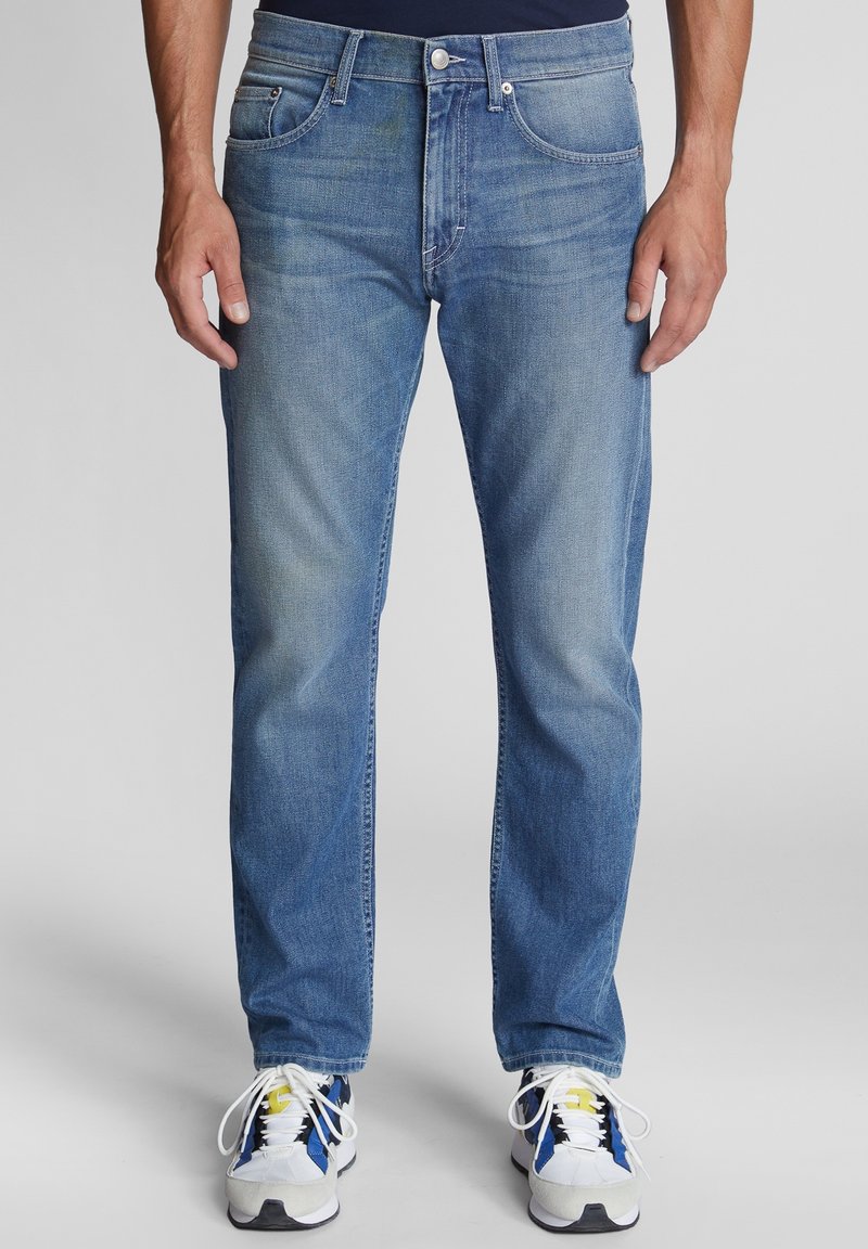 Homme portant un jean bleu moyen coupe droite et des baskets blanches avec des accents bleus et jaunes, debout devant un fond uni.