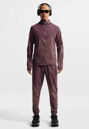 Uomo in piedi che indossa una tuta viola, occhiali da sole neri, cuffie over-ear e scarpe sportive nere su sfondo bianco.