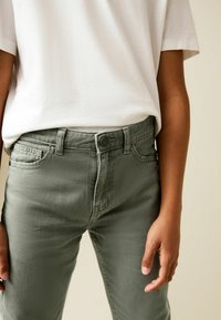 Zaļa denim džinsi ar šauru piegriezumu, ar klasisko piecu kabatu dizainu un pogu aizdari. Kombinēti ar vienkāršu baltu T-kreklu.