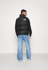 Black padded vest with high collar and logo on the back, worn over a black long-sleeve shirt. Blue wide-leg jeans and white sneakers.