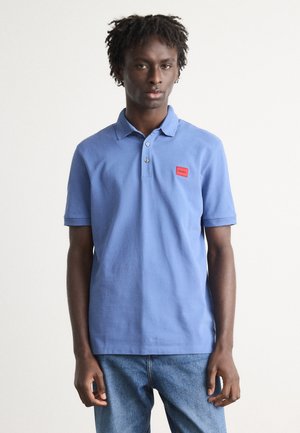 Polo bleu avec un col texturé, trois boutons et un écusson logo rouge. Porté avec un jean bleu clair, à manches courtes.