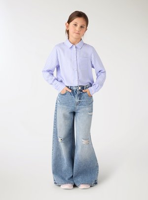 Ragazza giovane con le mani nelle tasche, indossa una camicia a righe blu e bianca e jeans a gamba larga usurati, in piedi su uno sfondo semplice.