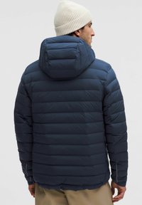 Tengerész kék puffdzseki nagy kapucnival, steppelt textúrával és elasztikus mandzsettával. Hátul vízszintes vonalak és lekerekített alj található.