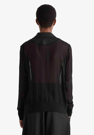 Persona con capelli corti e ricci che indossa una camicia nera trasparente a maniche lunghe vista da dietro su sfondo bianco.