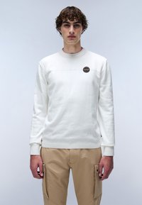 Sweatshirt blanc à manches longues, avec un col côtel�é et une couture horizontale. Présente un logo rond sur la poitrine. Porté avec un pantalon cargo beige.