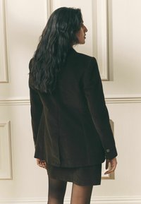 Barna kordbársony blézer, testhez simuló szabással, egyetlen hátsó sliccel és gombdísszel az ujjon. Egy hozzáillő szoknya fölött viselve.