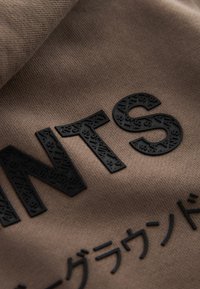 Tissu marron avec des lettres noires en relief "NTS" et des caractères japonais, texturé avec de plus petites lettres embossées à l’intérieur des grandes.