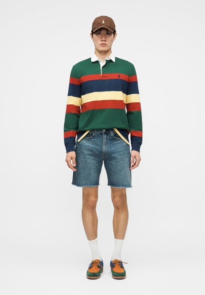 Άνδρας που φοράει καφέ καπέλο, μακρυμάνικο πόλο με πράσινες, κόκκινες, ναυτικές και κίτρινες ρίγες, τζιν σορτς, λευκές κάλτσες και πολύχρωμα παπούτσια με κορδόνια.