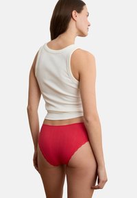 Frau trägt ein weißes ärmelloses Oberteil und rote Unterwäsche mit Spitzenbesatz, steht mit dem Rücken zur Kamera vor einem schlichten Hintergrund.