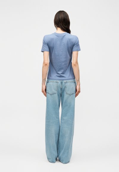 Γυναίκα με μακριά σκοτεινά μαλλιά που φορά κοντομάνικο μπλε πουκάμισο και φαρδιά ανοιχτό μπλε τζιν, στέκεται γυρισμένη με την πλάτη σε μονόχρωμο φόντο.