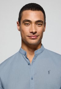 Ljusblå skjorta med knappar och mandarin-krage, tillverkad av ett strukturerat tyg. Har en broderad logotyp på bröstet.