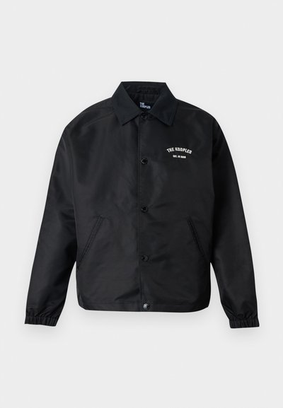 Μαύρο τζάκετ προπονητή με κουμπιά, μακριά μανίκια, ελαστικές μανσέτες, πλαϊνές τσέπες και λευκό κείμενο "THE KOOPLES EST. IN 2008" στο στήθος.