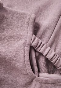 Detailní záběr na texturovanou mauve tkaninu s elastickým páskem a prošitým otvorem, ukazující záhyby látky a jemné detaily.