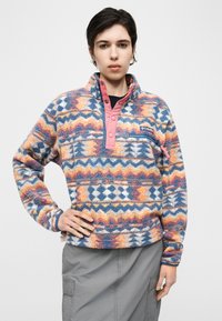 Γυναίκα που φοράει πολύχρωμη φλις μπλούζα με μοτίβο και ροζ κουμπιά με πατιλέτα και γκρι φούστα, στέκεται με το ένα χέρι στη μέση.