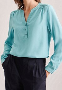 Blusa turchese con scollatura a V, chiusura a cinque bottoni e maniche lunghe; tessuto liscio; dettagli plissettati sulle spalle; abbinata a pantaloni scuri.